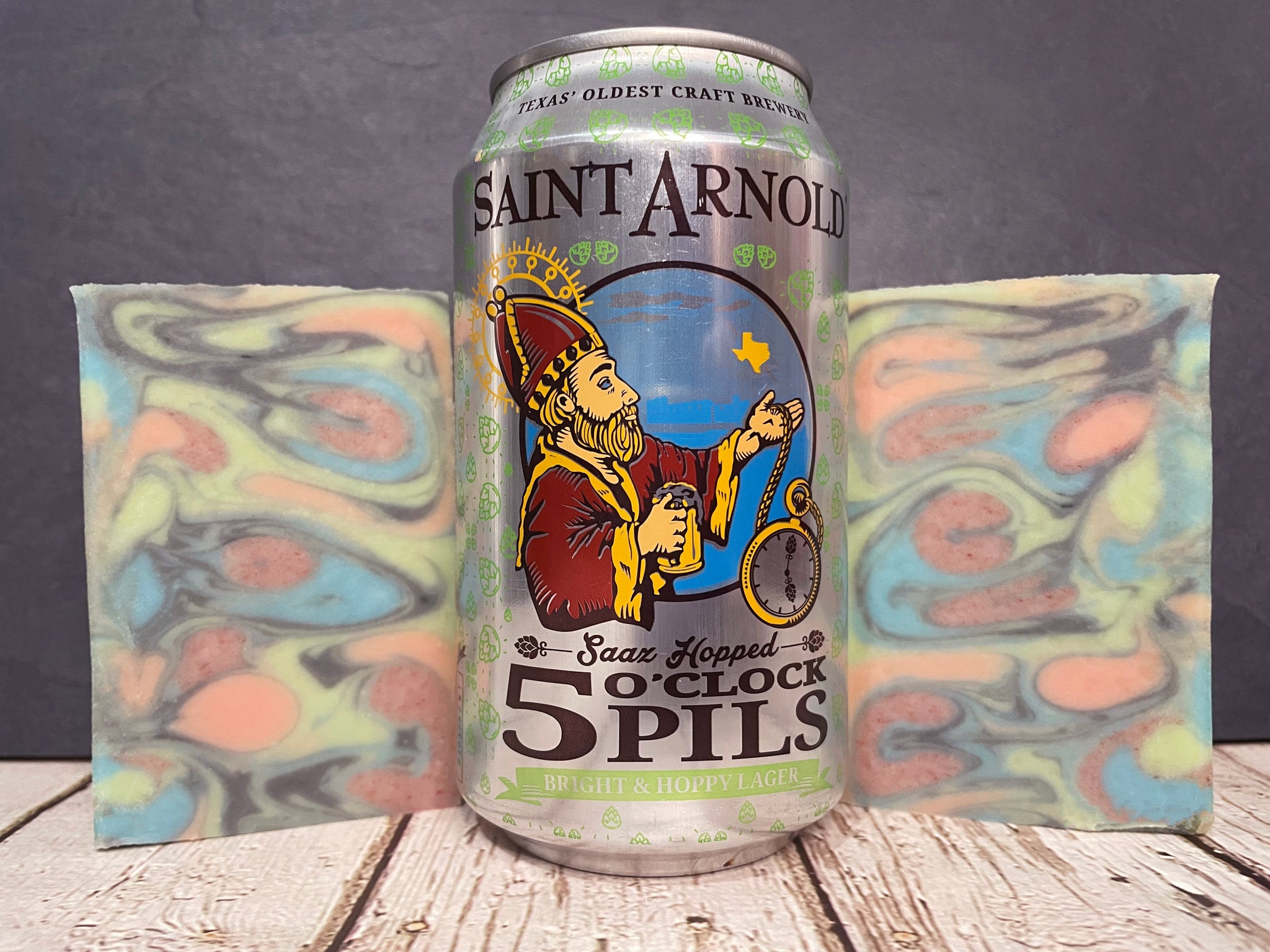 Two swirl beer soaps made with 5 oclock pils beside a can of Saint Arnold Brewing Company 5 o'clock Pils on a wooden surface with a dark background