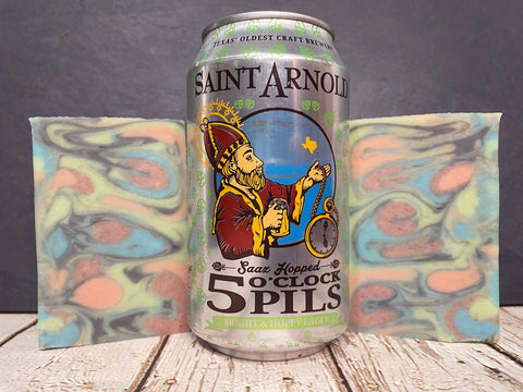 Two swirl beer soaps made with 5 oclock pils beside a can of Saint Arnold Brewing Company 5 o'clock Pils on a wooden surface with a dark background
