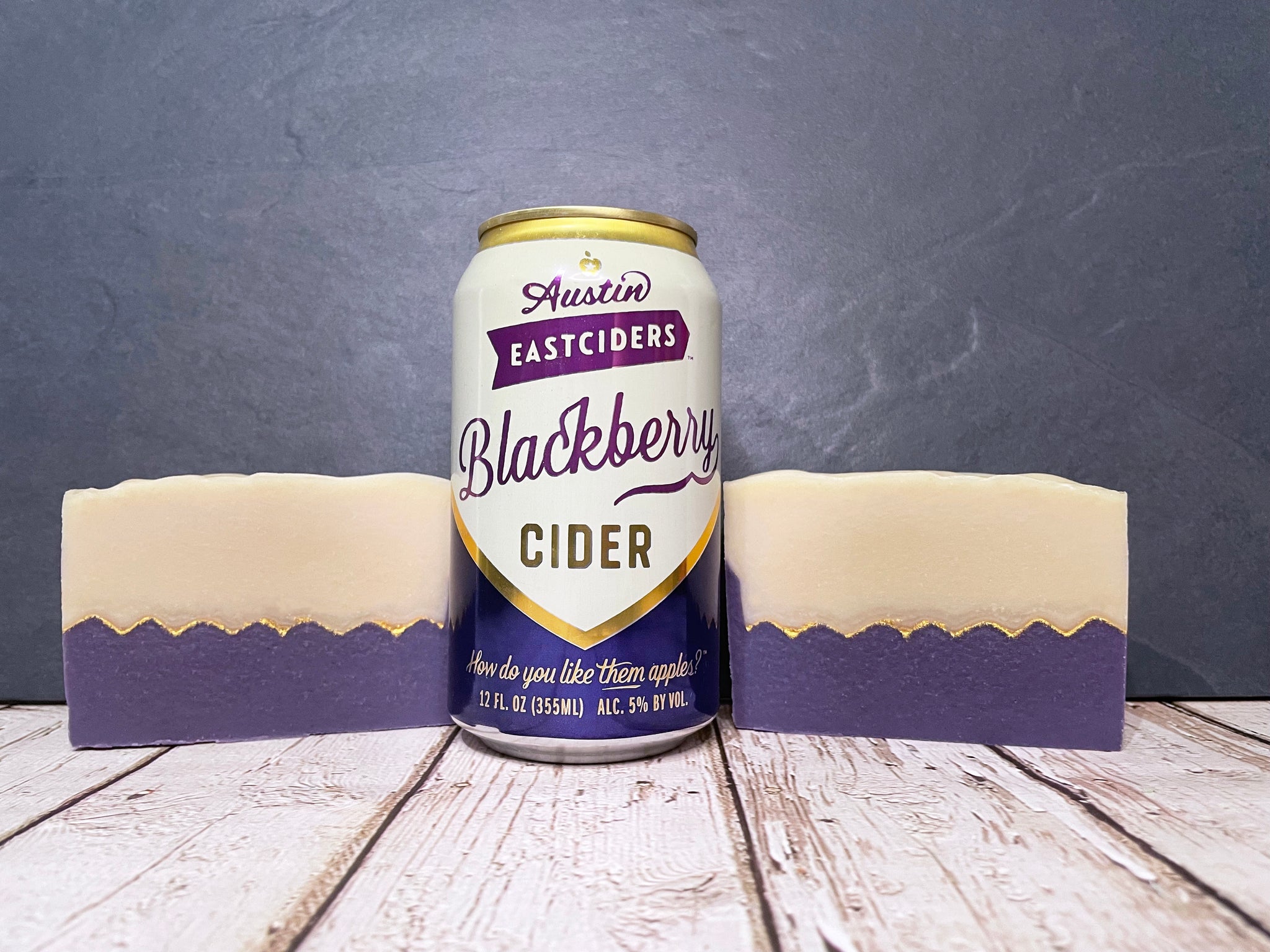 Austin Eastciders Blackberry Cider can between two bars of purple and cream cider soap on a wooden surface with a gray background
