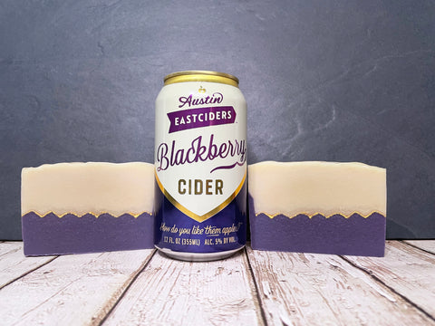 Austin Eastciders Blackberry Cider can between two bars of purple and cream cider soap on a wooden surface with a gray background