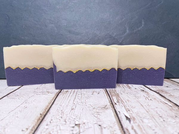 Three bars of cider soap with gold mica line and purple and white layers on a wooden surface.