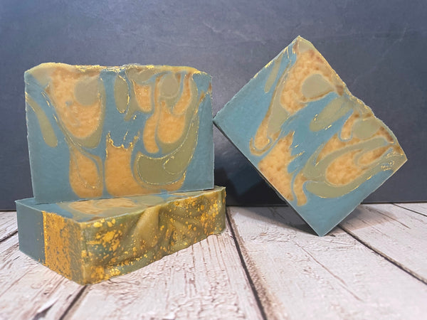 Two blue and gold bars of beer soap with a marbled design on a wooden surface against a dark background