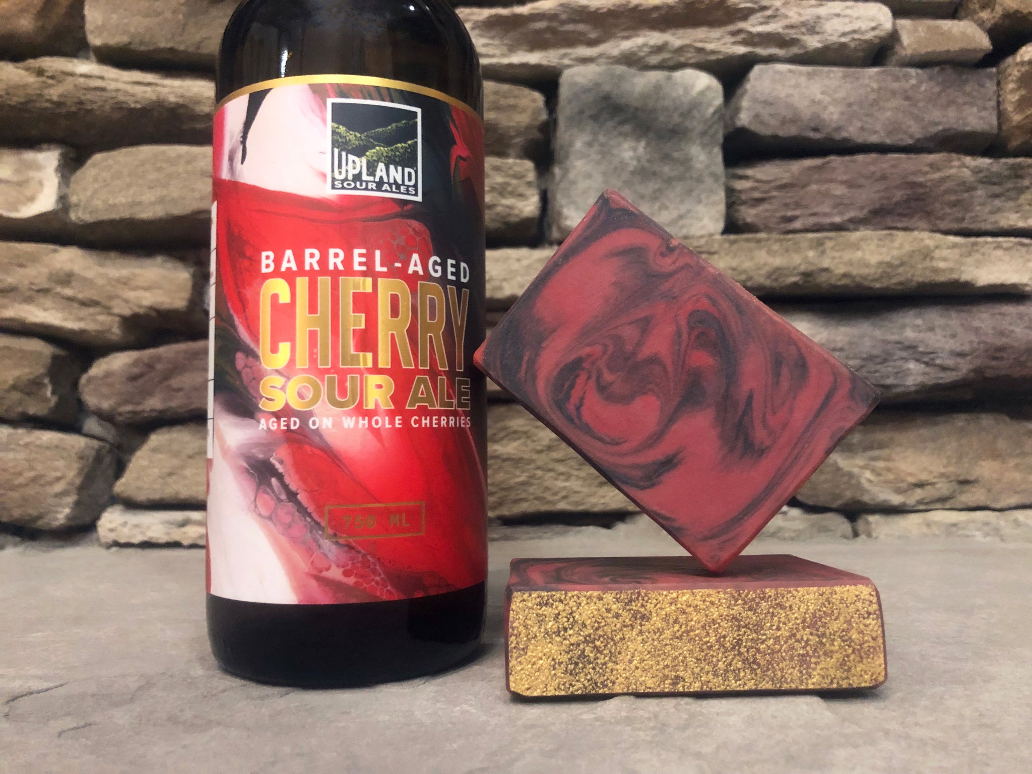 Bottle of Upland Barrel-Aged Cherry Sour Ale beside two bars of red and black beer soap with activated charcoal on a stone surface.