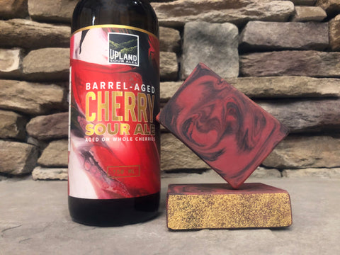 Bottle of Upland Barrel-Aged Cherry Sour Ale beside two bars of red and black beer soap with activated charcoal on a stone surface.