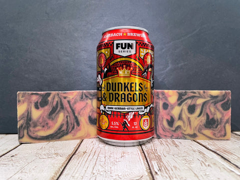 Can of Dunkels & Dragons beer by Karbach Brewing Company between two marbled gold red and black beer soap bars on a wooden surface with a dark background.