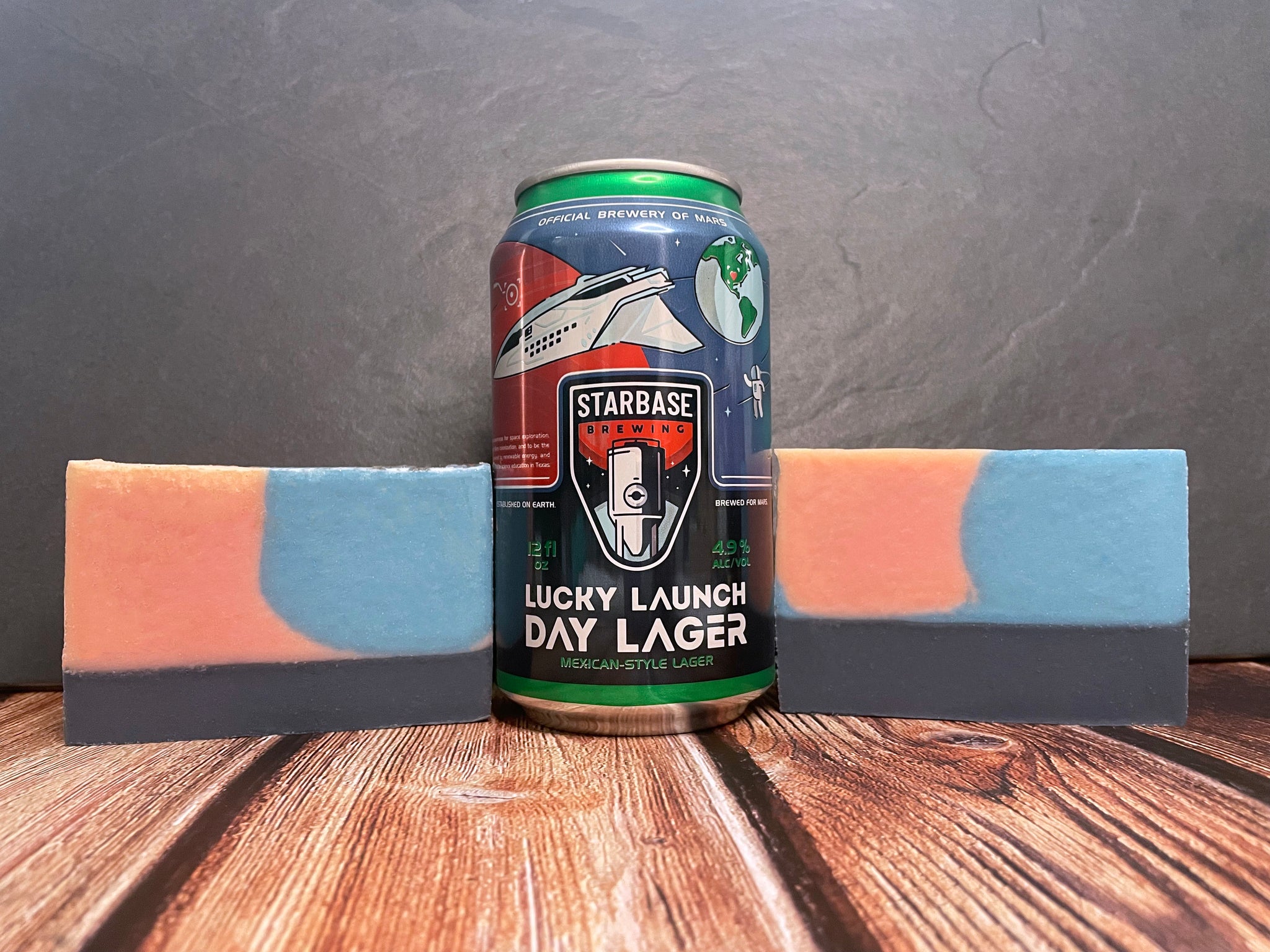 two red black and blue beer soap bars by spunkndisorderly beer soaps alongside a starbase brewing lucky launch day lager beer can on a wooden surface