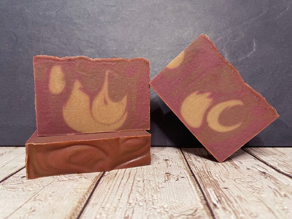 Two beer soap bars with maroon and gold drop swirl design on a wooden surface against a dark background