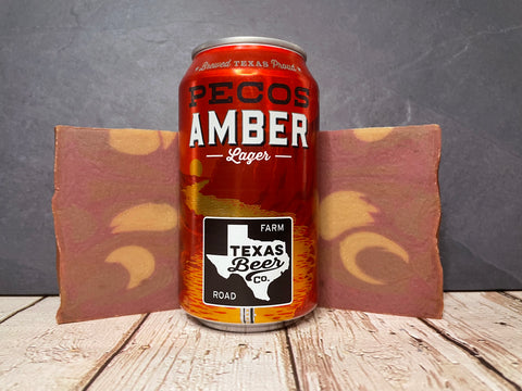 Pecos Amber Lager Texas Beer Co. can between two maroon and gold bars of beer soap on a wooden surface
