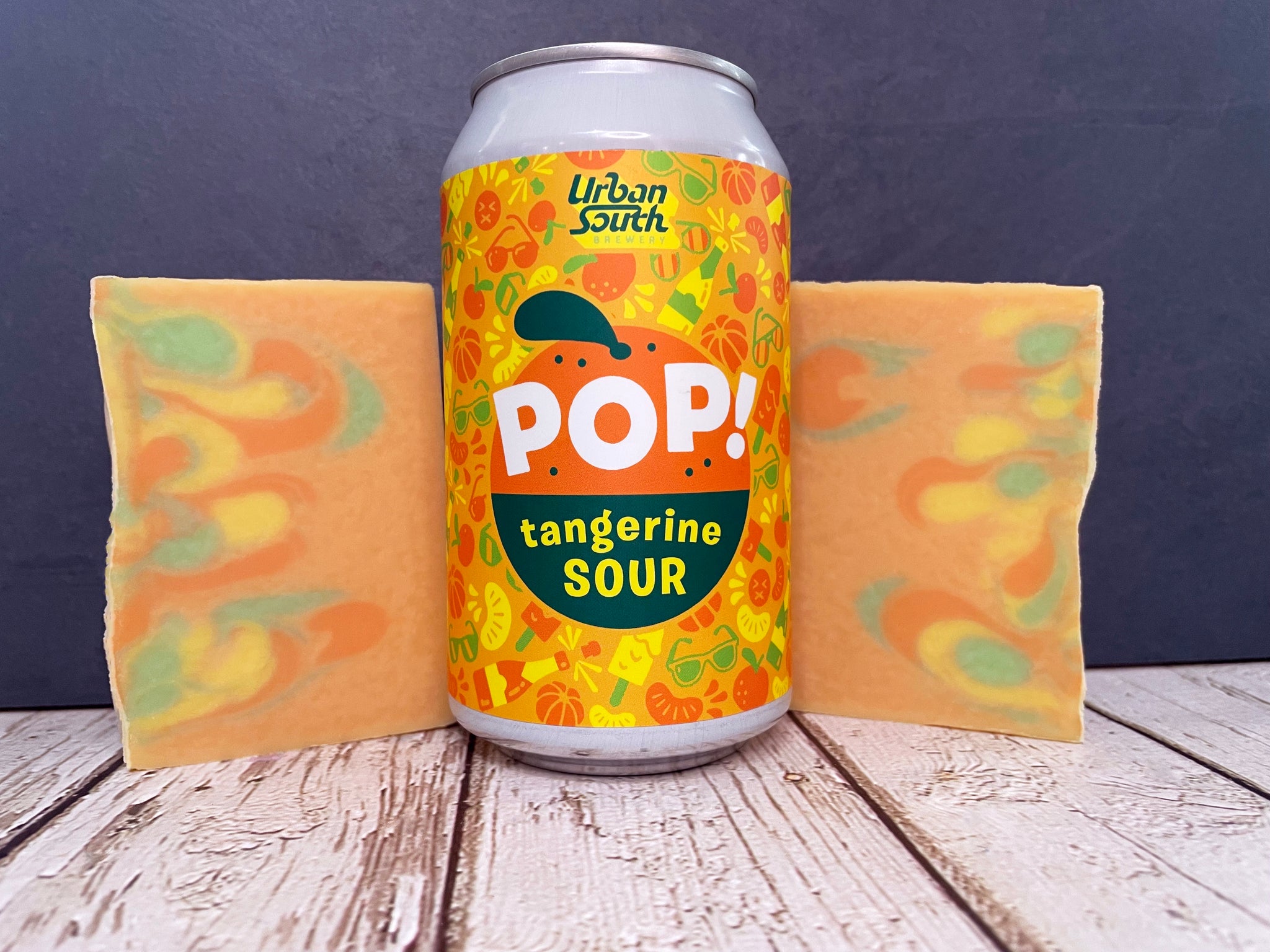 pop tangerine sour urban south brewery beer can between two bars of orange yellow and green marbled beer soap on a wooden surface with stone background