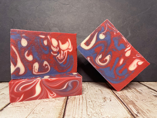 Three marbled beer soap bars in red, blue, and white on a wooden surface with a dark background.