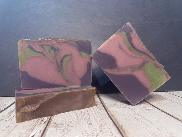 Three bars of beer soap with a purple and green swirl pattern on a wooden surface with a dark background.