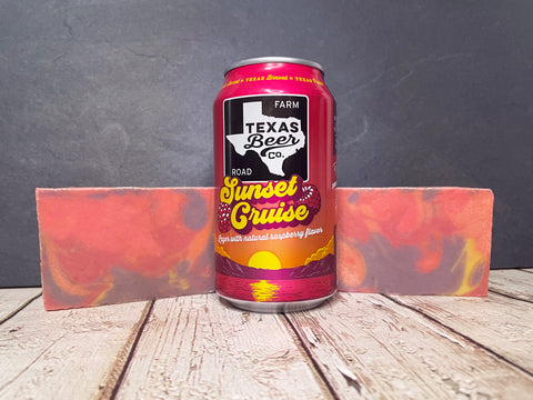 Can of Texas Beer Co. Sunset Cruise beer between two purple pink and yellow swirl beer soap on a wooden surface with a dark background