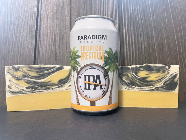 Paradigm Brewing Tropical Precision IPA can on a wooden surface with two yellow white and black beer soap bars with activated charcoal