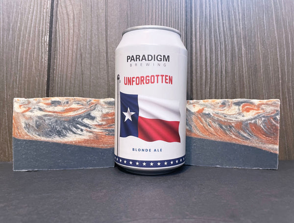 Paradigm Brewing Unforgotten Blonde Ale beer can between two bars of red white and blue beer soap on a stone surface