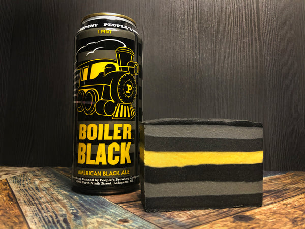 can of boiler black American black ale  from people's brewing company beside a bar of black grey and yellow striped beer soap on a wooden surface with a black background